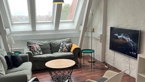 Ein modernes Wohnzimmer mit hohen Decken, Dachfenstern, einem runden Holztisch, einer Couch, zwei Sesseln und einem an der Wand befestigten Flachbildfernseher.