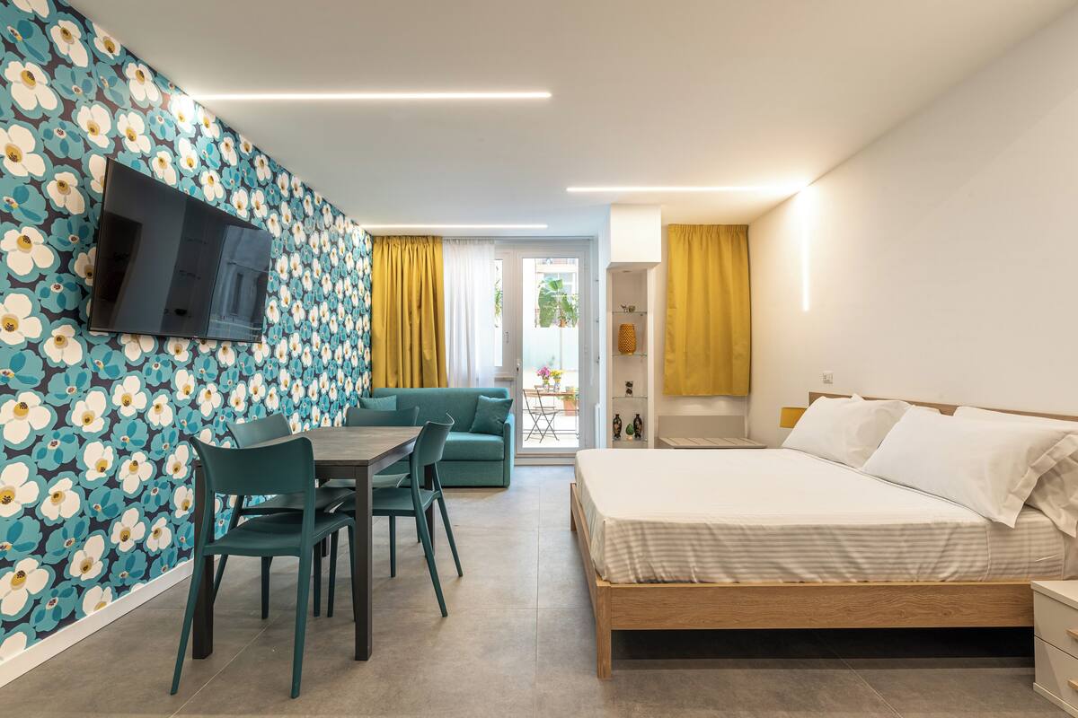 Ein modernes Zimmer mit einem Bett, einem Esstisch mit Stühlen, einem an der Wand befestigten Fernseher und einer Wandtapete mit Muster.