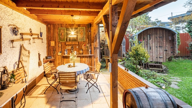 Un intérieur rustique avec des éléments en bois, notamment une table ronde et des chaises, un plafond en bois et une vue sur un jardin agrémenté d’un abri de jardin en bois.
