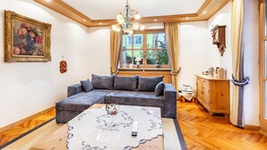 Un salon comprenant un canapé d’angle, une table basse, un lustre, une fenêtre avec des rideaux et un meuble en bois.