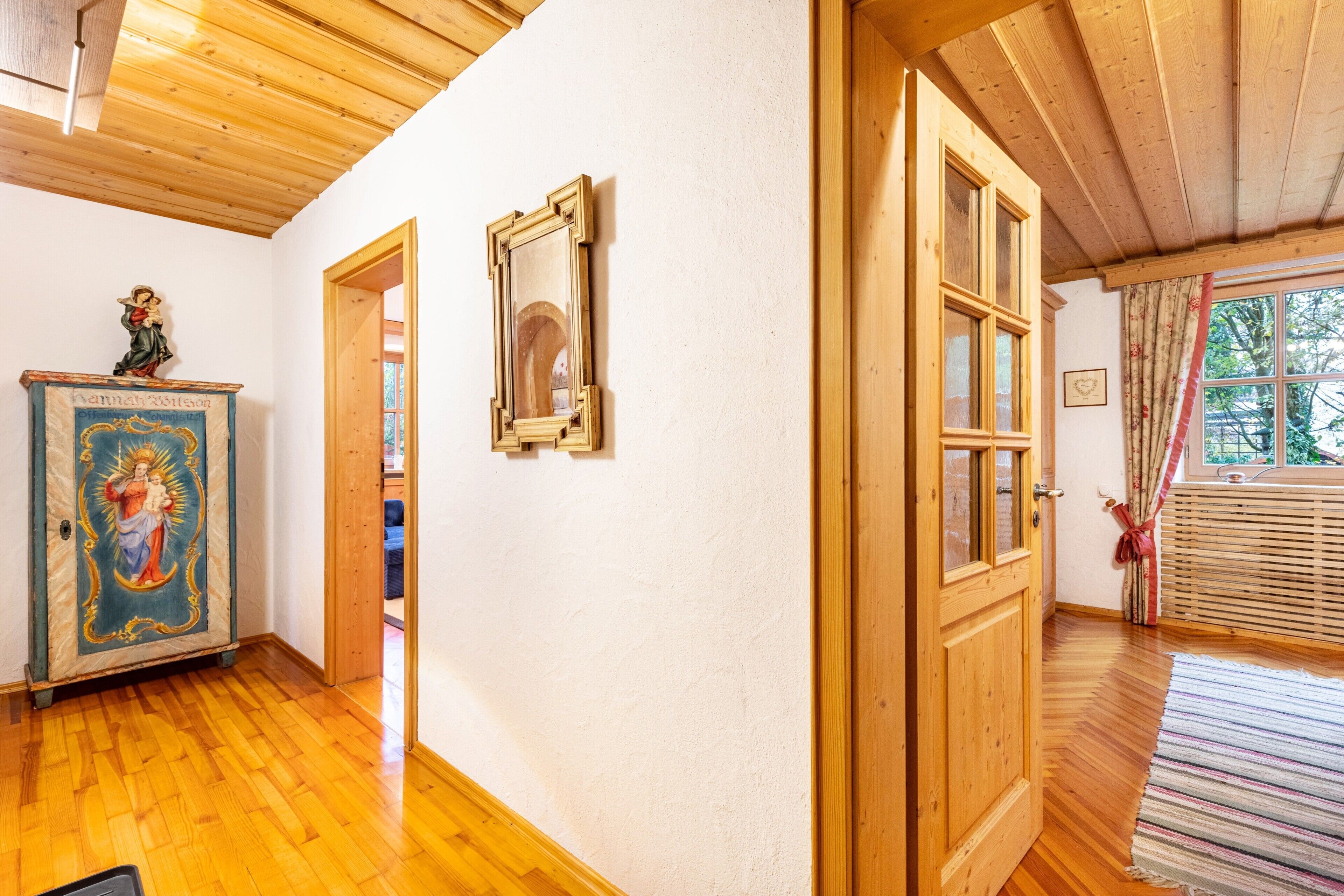 Une pièce avec un plancher en bois, un plafond en bois, une porte, une fenêtre avec des rideaux et un mur orné d’un tableau encadré.