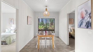 Un couloir avec une table à manger dressée pour quatre personnes, une grande fenêtre donnant sur une forêt, et un lit visible au fond.
