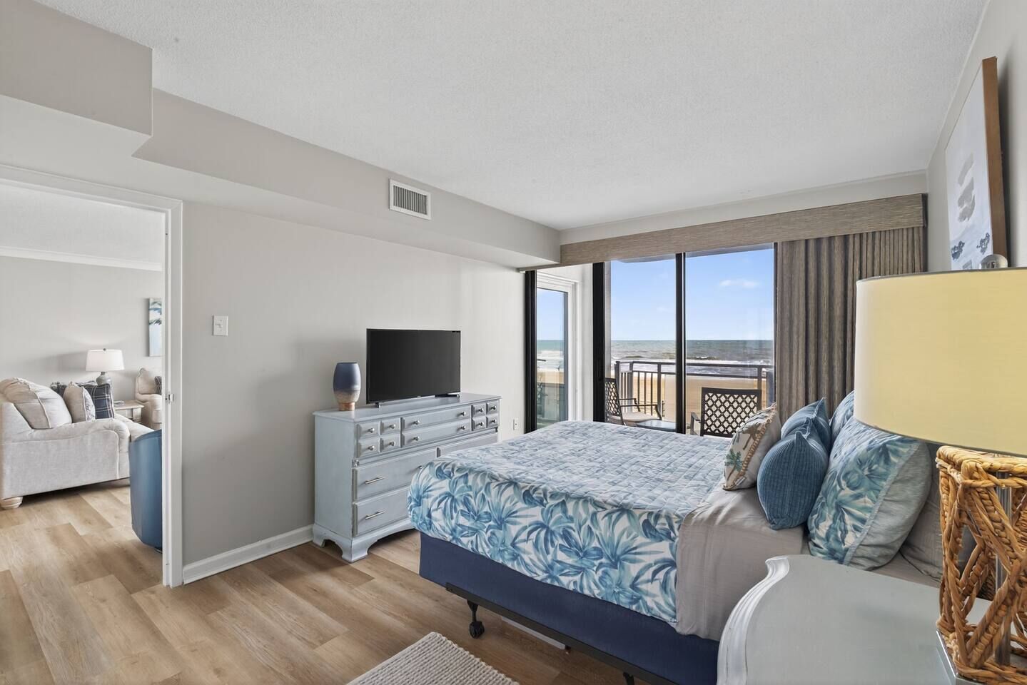 A bedroom with a bed, dresser, TV, and a balcony with a view of the beach.