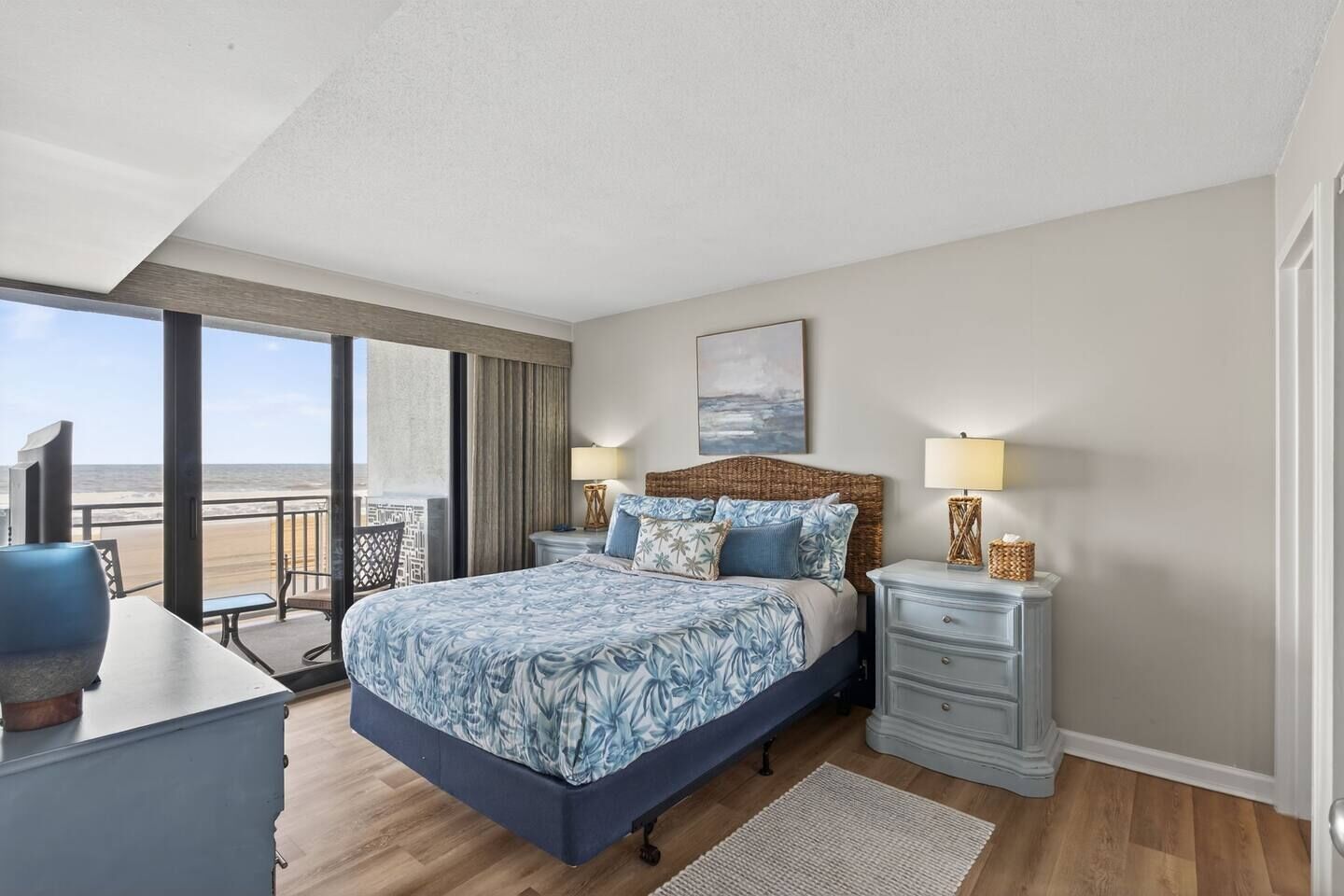 A bedroom with a large bed, a nightstand, a dresser, and a view of the beach.