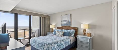 A bedroom with a large bed, a nightstand, a dresser, and a view of the beach.