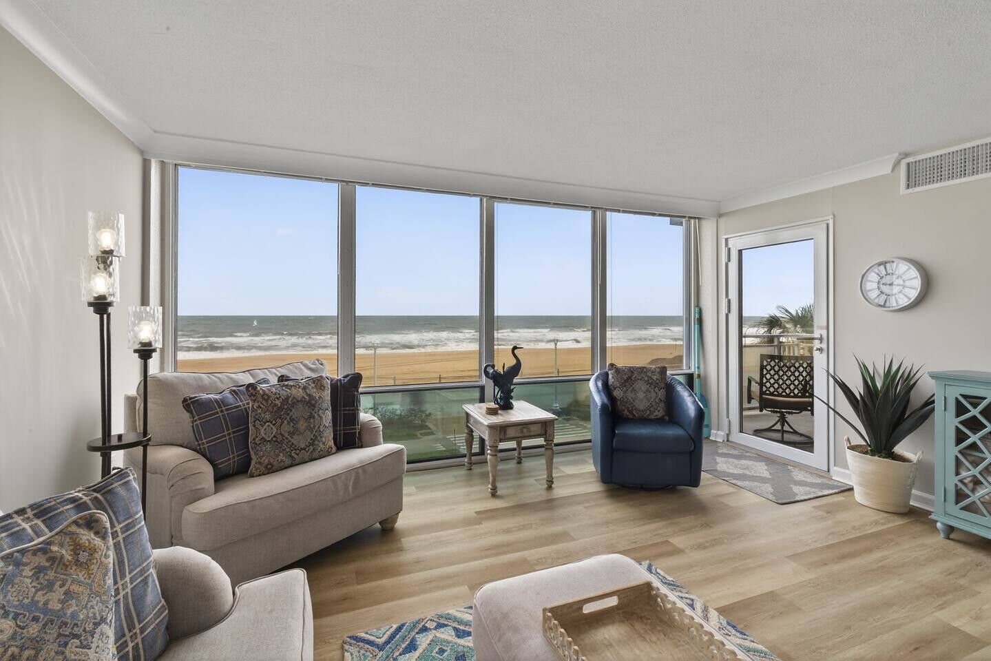 A spacious living room with a large view of the beach, a blue armchair, a beige sofa, and a small table with decorative items.