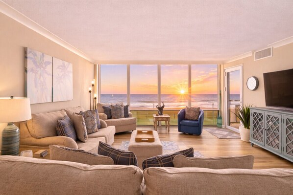 Ein Wohnzimmer mit einer Couch, Sesseln, einem Couchtisch und einem Fernseher. Ein großes Fenster bietet Blick auf den Ozean und einen Sonnenuntergang.