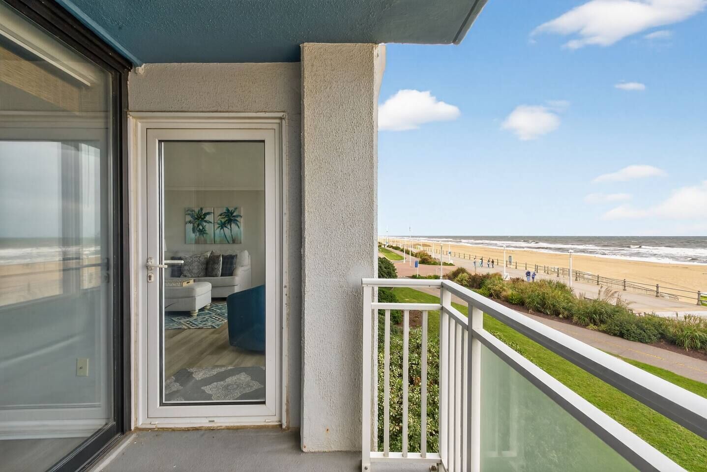 A balcony with a view of the beach, a glass door leading to a living area with a sofa and a coffee table, and a railing with greenery.
