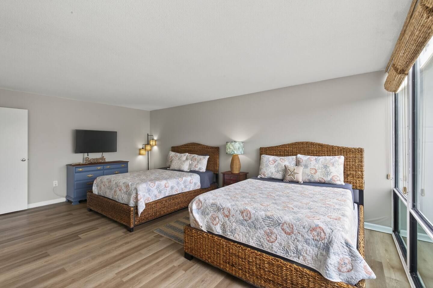 A bedroom with two wicker beds, a blue dresser, a flat-screen TV, and a large window.