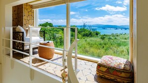 Ein Balkon mit einer weißen Couch, einem gewebten Korb und einem farbenfrohen Hocker. Ein großes Fenster bietet Blick auf Grünflächen und ein Gewässer.