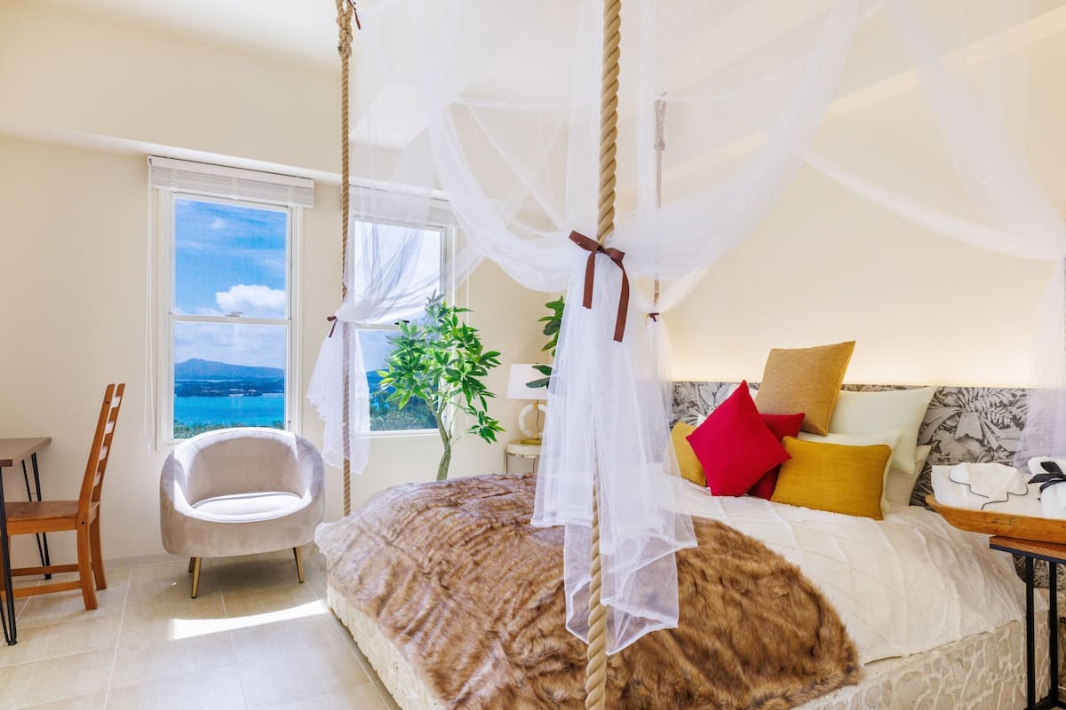 Een slaapkamer met een hemelbed, een stoel, een plant en uitzicht op zee door een raam.
