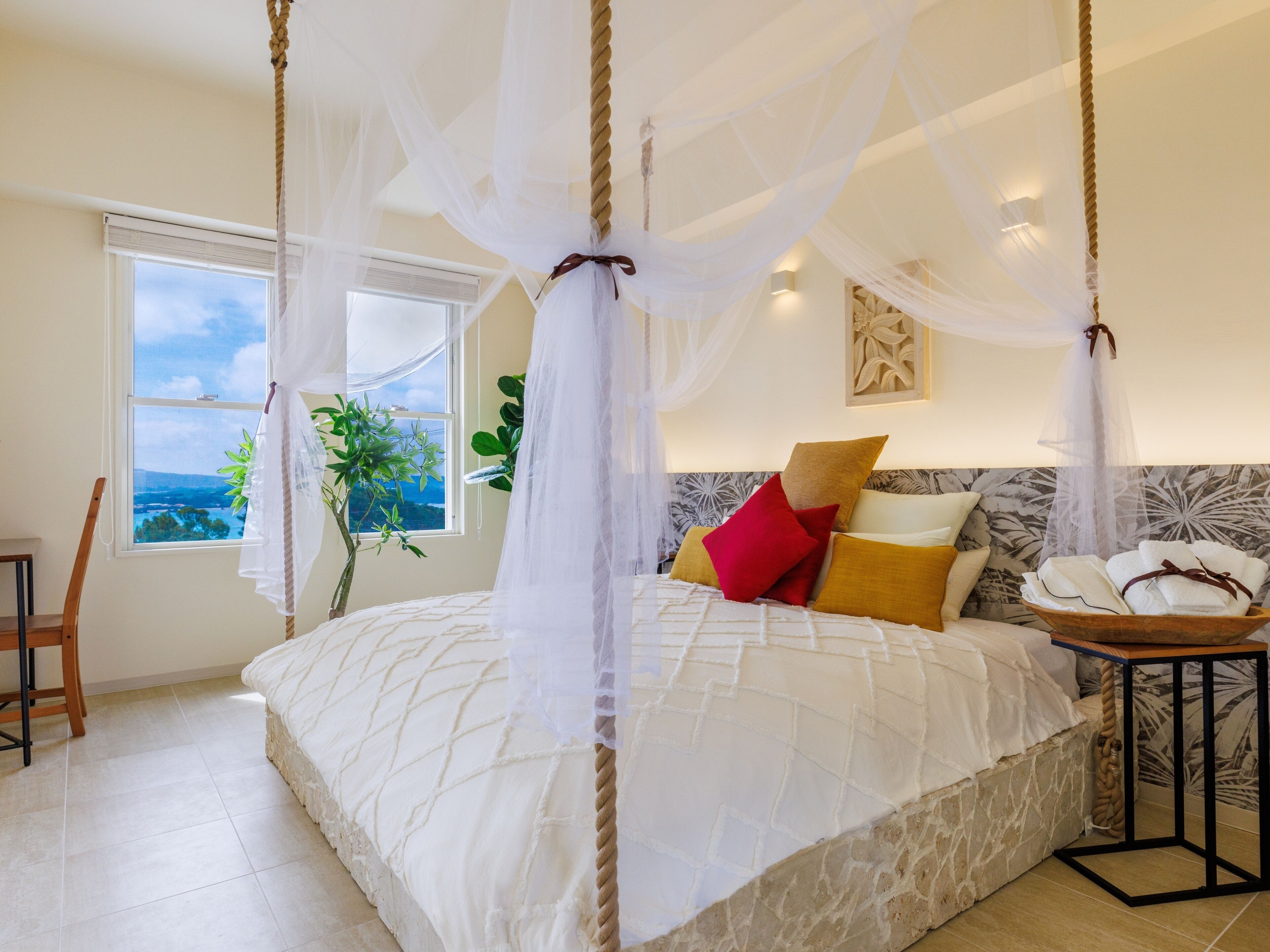 A bedroom with a four-poster canopy bed, a view of the outdoors through a window, and a bedside table with towels.