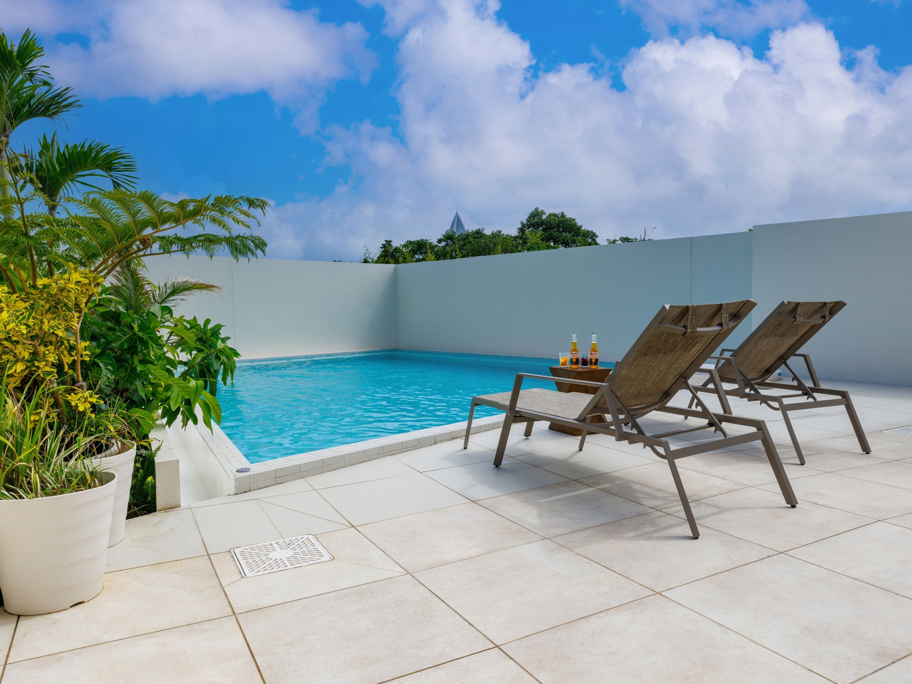 A poolside area with two lounge chairs and a small table with drinks, surrounded by potted plants and a clear blue sky.