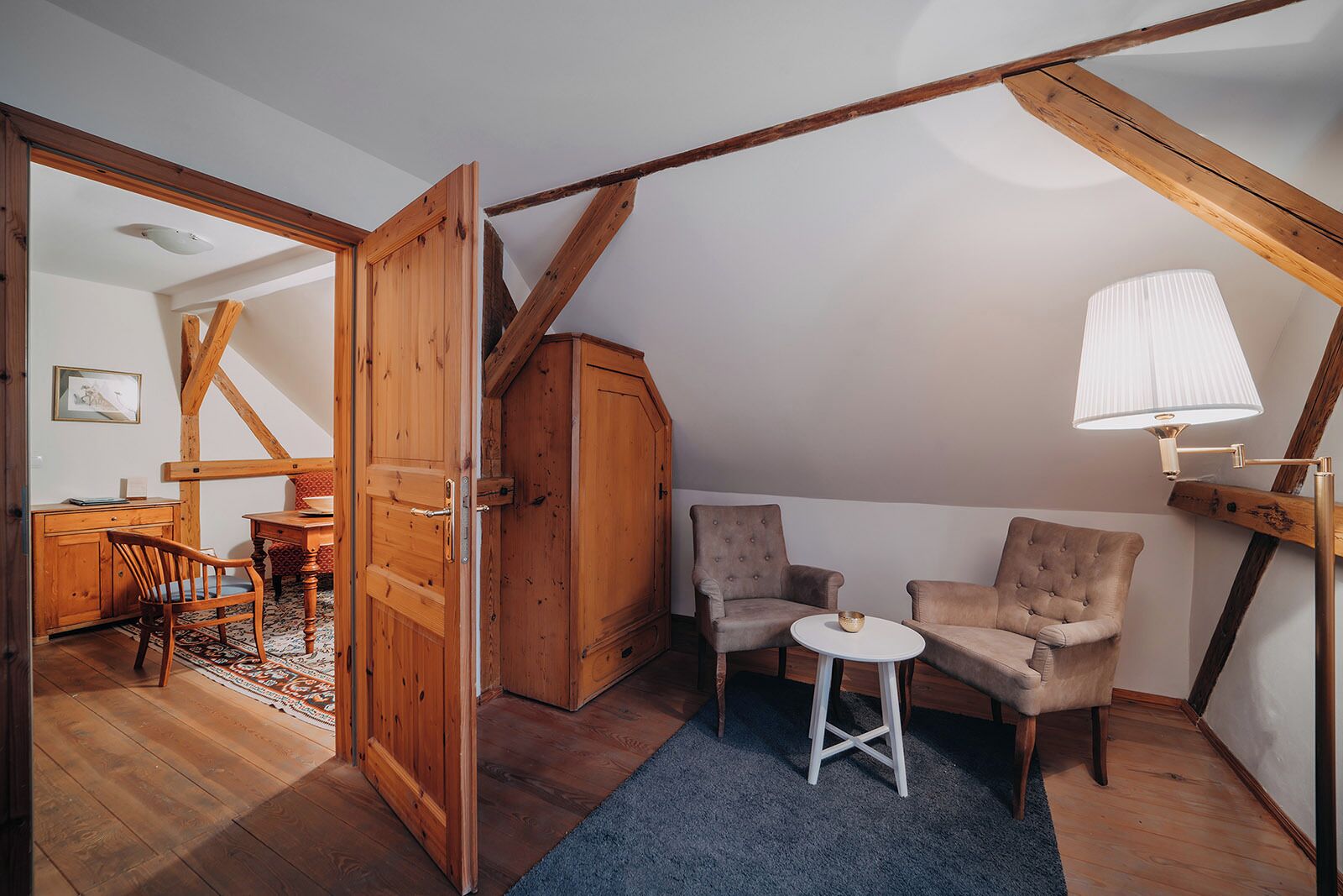 Una habitación acogedora en el desván, con vigas de madera a la vista, un armario de madera, dos sillones, una mesita redonda y una lámpara de pie.