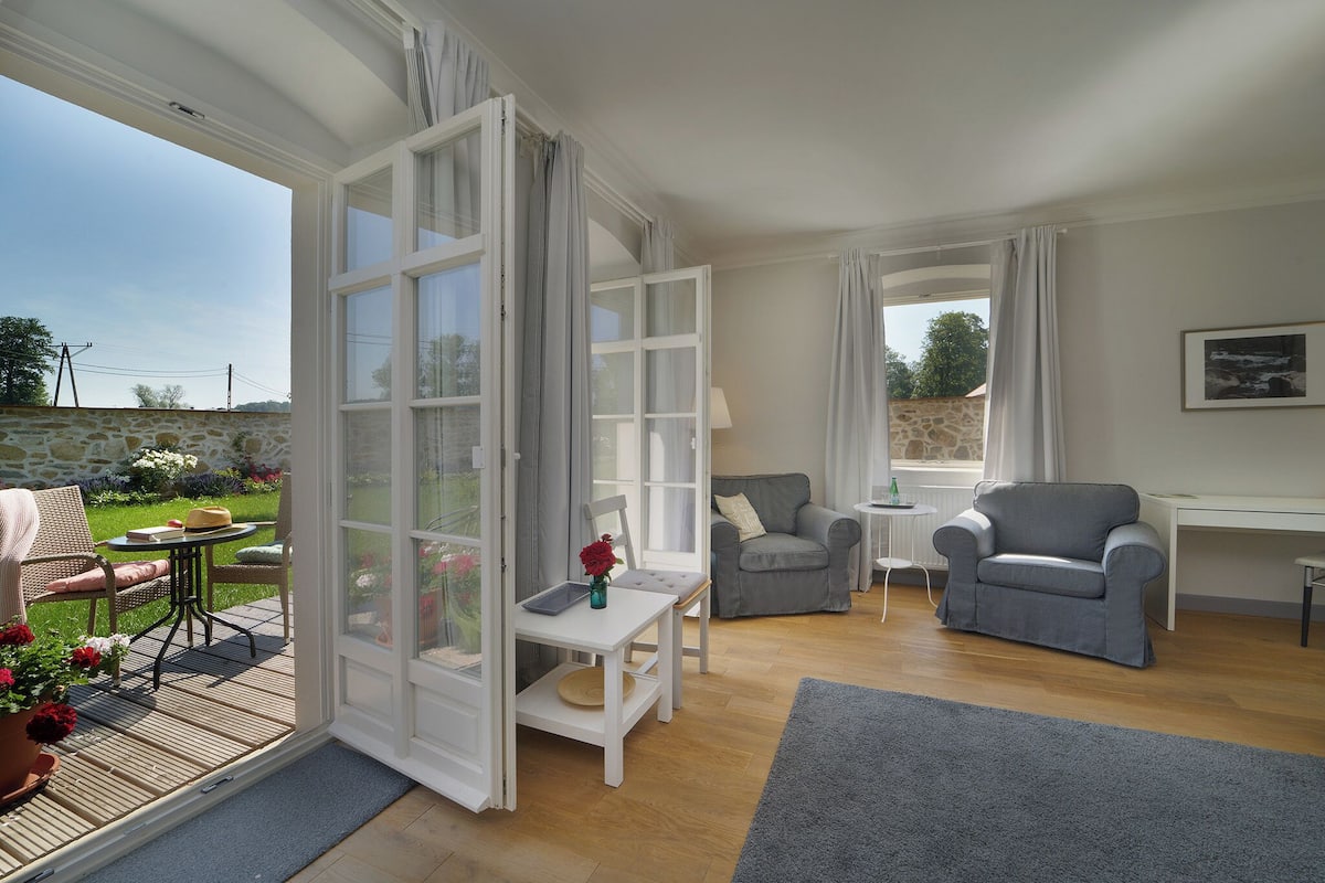 Amplia sala de estar con un gran ventanal que da a un jardín, un sillón gris, una mesita con un jarrón de flores y un sofá blanco.