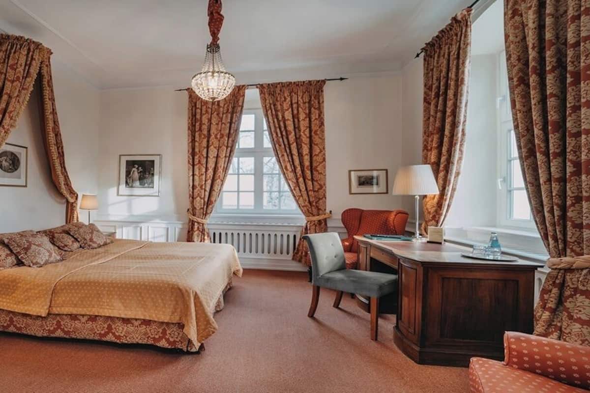 Un dormitorio con una cama grande, un escritorio y dos sillas. Hay cortinas en las ventanas y una araña colgando del techo.