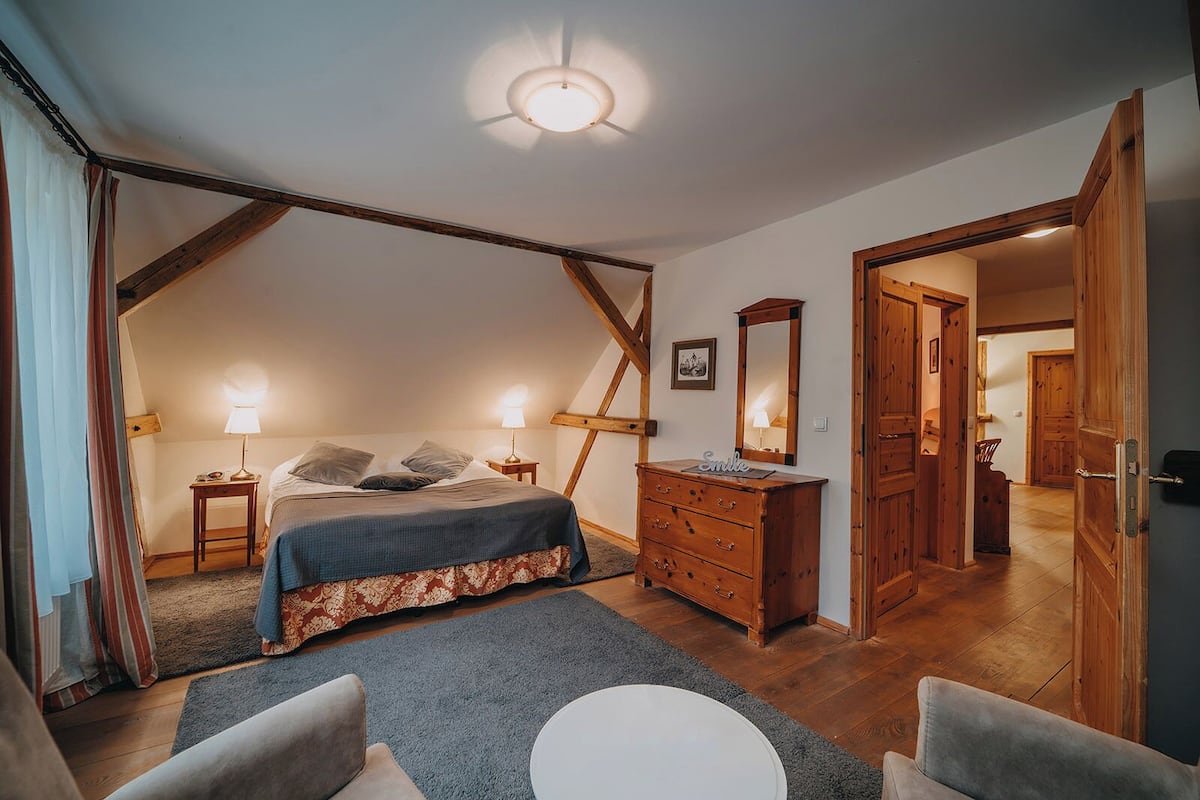 Un dormitorio acogedor con techo inclinado, una cama con colcha estampada, un aparador, un espejo y una puerta que da a otra habitación.