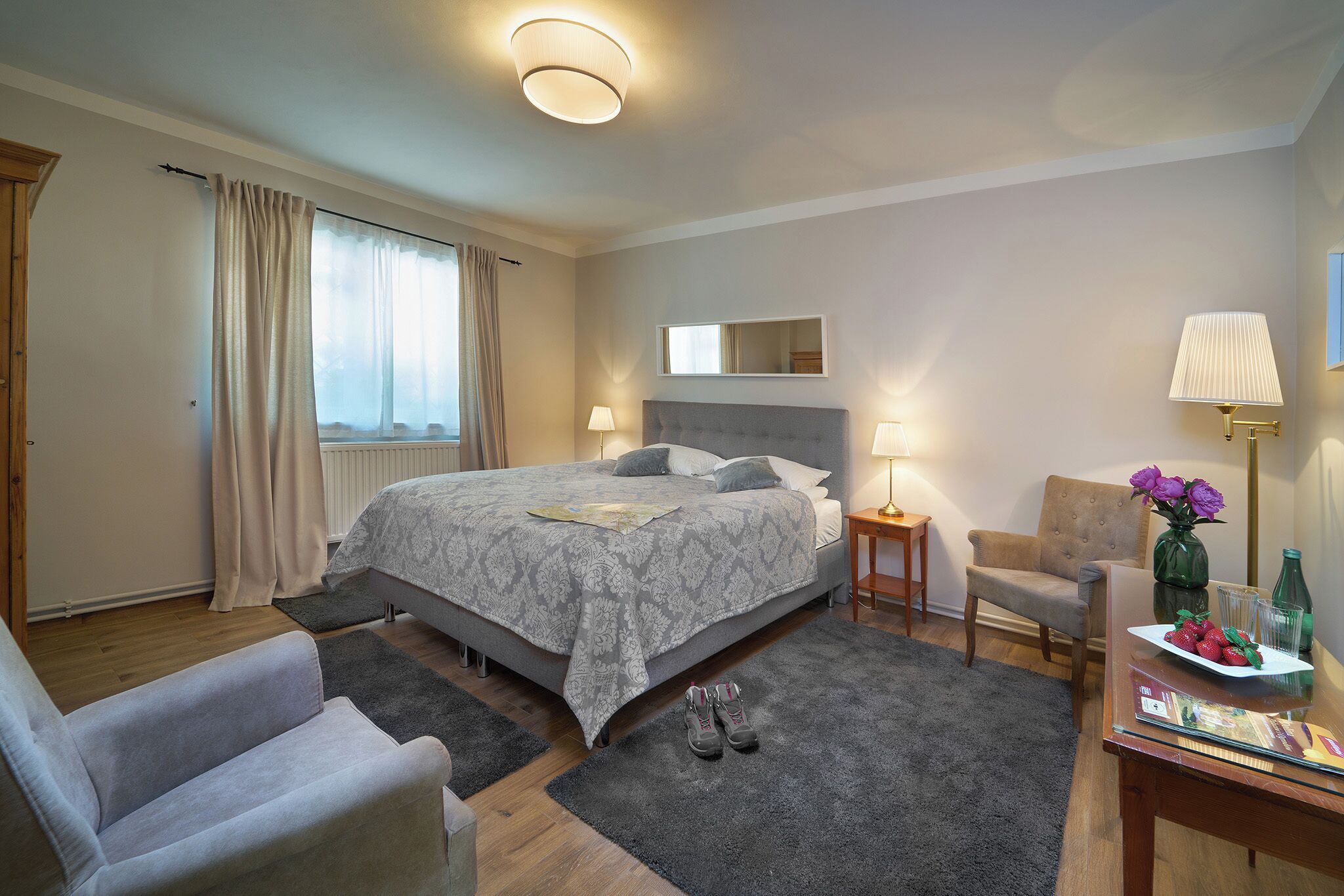 Habitación de hotel impecable con una cama grande, una zona de estar con una silla, una mesita con un florero y vasos, y una ventana con cortinas.