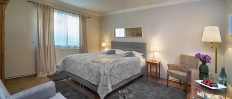 Habitación de hotel impecable con una cama grande, una zona de estar con una silla, una mesita con un florero y vasos, y una ventana con cortinas.