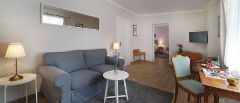A modern living room with a grey sofa, a small white table, a wooden dining table with chairs, a TV mounted on the wall, and a mirror above the sofa.