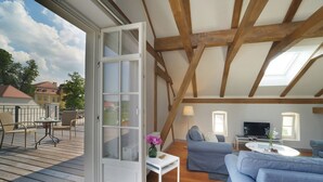 Eine offene Tür zu einem Balkon mit Holzterrasse, einer Couch, einem Fernseher und einem Tisch mit Blumenvase.