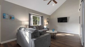 Un salon avec un canapé gris, une table basse ronde, un ventilateur de plafond, une fenêtre avec des rideaux et un téléviseur à écran plat fixé au mur.
