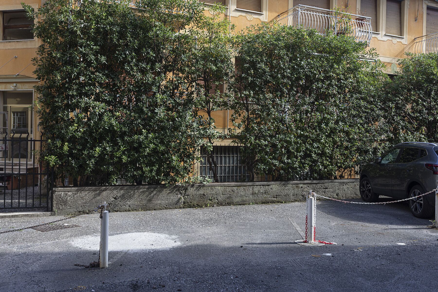 A parking area with a chain-locked car, a building with balconies, and dense foliage.