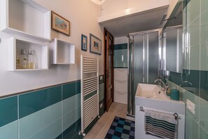 A bathroom with a shower, sink, and towel rack. There are framed pictures on the wall and a shelf with toiletries.