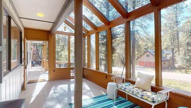 A wooden cabin with large windows, a porch with a swing, and a view of a forest and a red building in the distance. - Pet Friendly | Monte Verde Lake | Fireplace | Deer Casa (Angel Fire)
