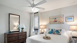 Una camera da letto con un letto grande, una cassettiera, una ventola a soffitto, una finestra con tende e una parete con un cartello e un quadro incorniciato.