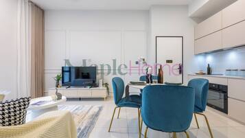 A modern living room with a TV, dining table, and chairs. There is a kitchen area with appliances and cabinets.