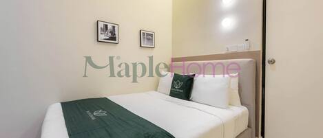 A neatly made bed with a green blanket and a pillow with a logo, a wall-mounted picture frame, and a door with a light switch.