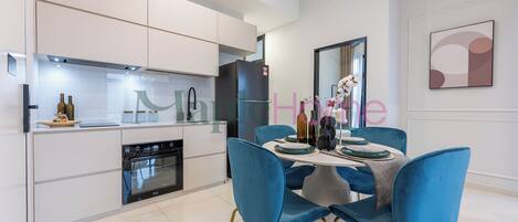 A modern kitchen with white cabinets, a black refrigerator, and a built-in oven. There is a dining area with blue chairs and a white table.