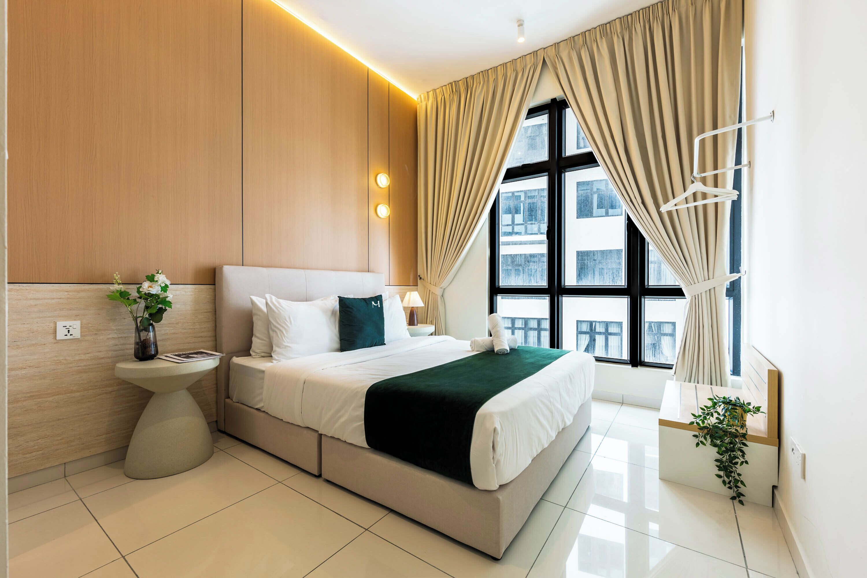 Habitación de hotel moderna con una cama grande, una mesita de noche, una lámpara y vistas de la ciudad a través de amplios ventanales.