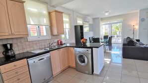 A modern kitchen with a dishwasher, washing machine, and coffee maker. There is a dining area with a table and chairs, and a living room with a sofa. - Aurora Villa Protaras - Private Pool Wifi (Paralimni)