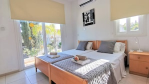 A bedroom with two beds, a nightstand, a bedside lamp, and a window with a view of greenery. - Aurora Villa Protaras - Private Pool Wifi (Paralimni)