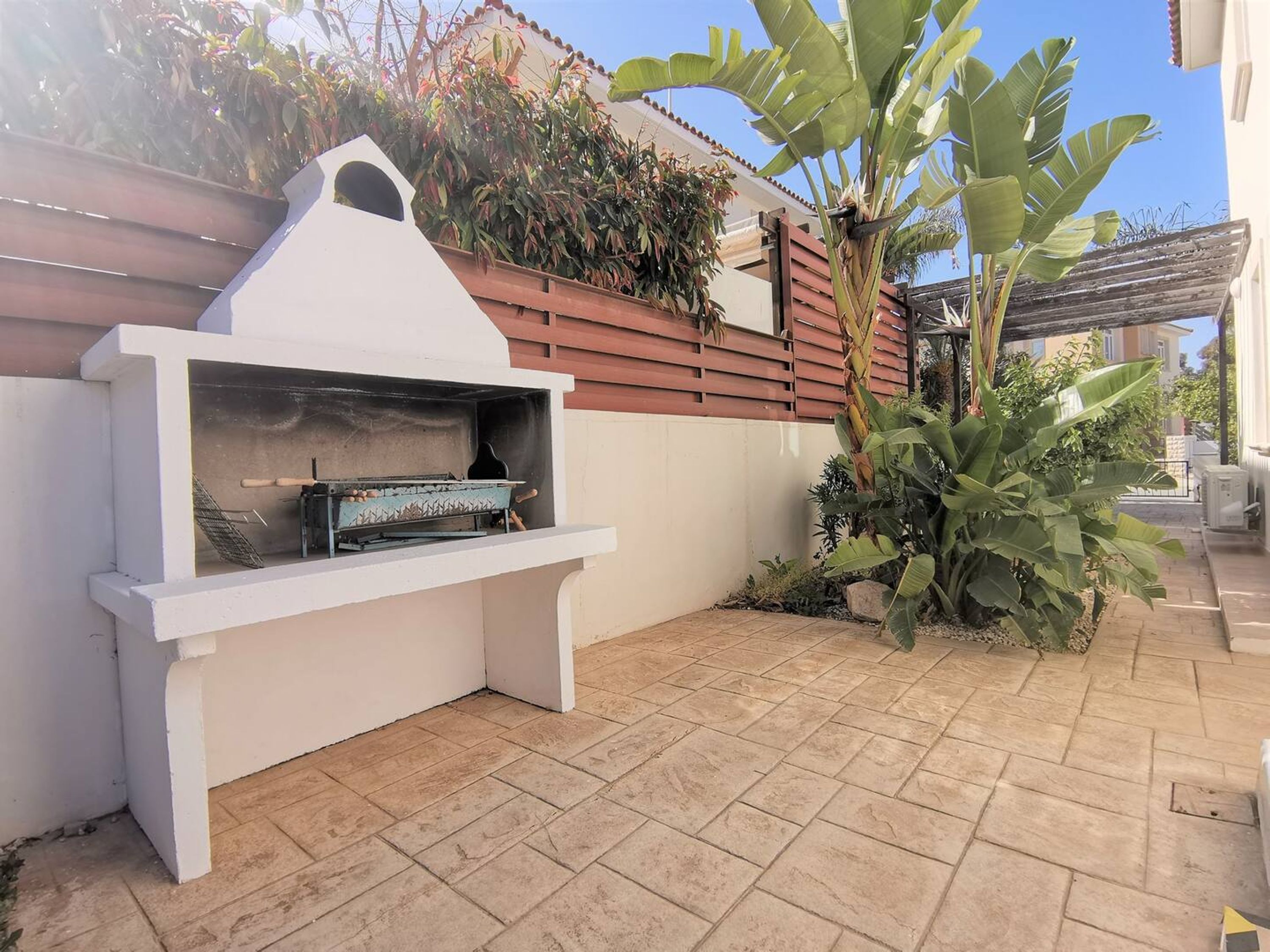Un barbecue extérieur blanc, doté d’une cheminée, une terrasse carrelée et un grand bananier.