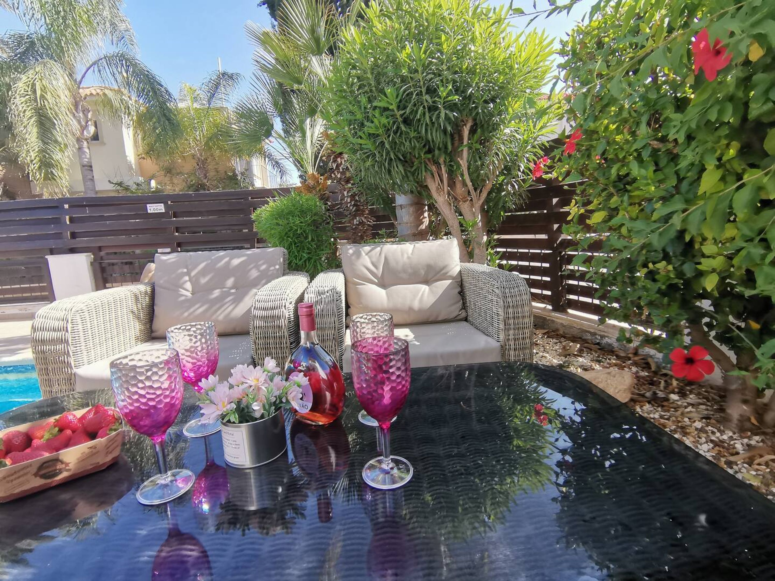 Un espace aménagé au bord de la piscine, avec des meubles en osier, une table en verre et divers objets décoratifs, notamment des fleurs et une bouteille.