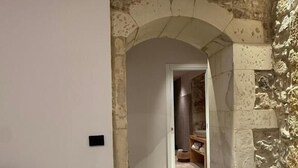 Un couloir avec une arche en pierre menant à une chambre. On y trouve deux lanternes blanches posées sur le sol, un banc blanc et une petite table sur laquelle se trouve un objet noir.