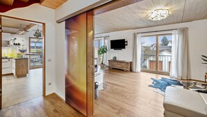 Amplia sala de estar con puerta corredera que da a una cocina, un televisor de pantalla plana montado en la pared y un gran ventanal con cortinas.