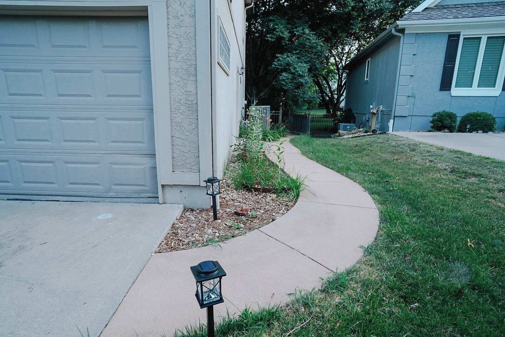 Une allée menant à une résidence, avec un trottoir incurvé, une porte de garage et un petit jardin agrémenté d’une lanterne.