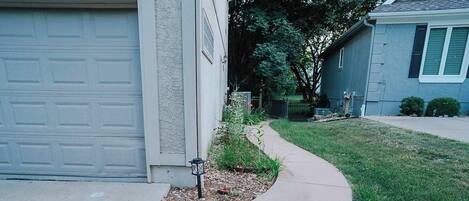 Une allée menant à une résidence, avec un trottoir incurvé, une porte de garage et un petit jardin agrémenté d’une lanterne.