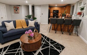 A living room with a sofa, coffee table, and chairs, a kitchen with wooden cabinets and a dining area with bar stools.
