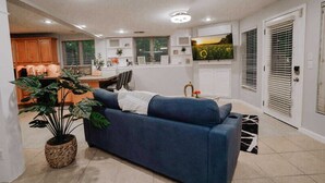 A living room with a sofa, a potted plant, and a kitchen in the background.