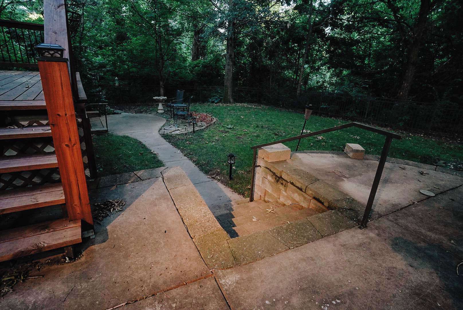 Une terrasse en bois avec un escalier en pierre menant à une zone herbeuse où se trouvent un banc et un abreuvoir à oiseaux.