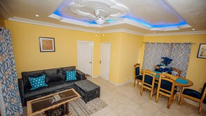 A living room with a black sofa, a wooden coffee table, and a dining area with wooden chairs and a table. - Bethel Suites Hotel (Accra)