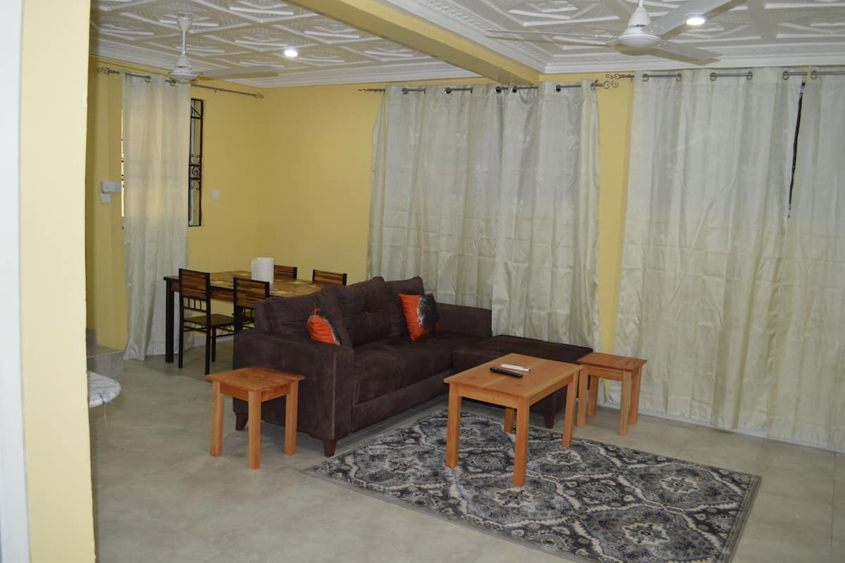 A living room with a brown sofa, two wooden side tables, a patterned rug, a dining table with chairs, and a ceiling fan.