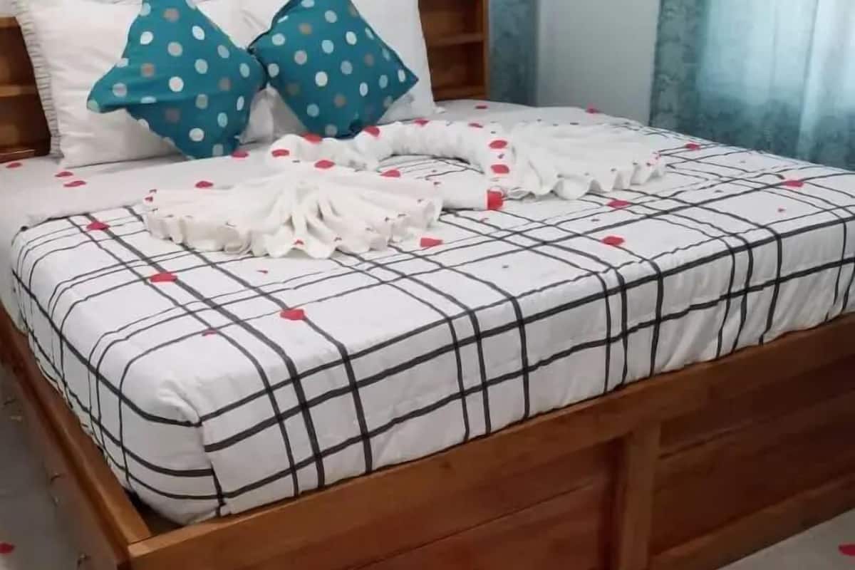 A bed with a plaid bedspread, two polka-dotted pillows, and a heart-shaped arrangement of rose petals on the floor.