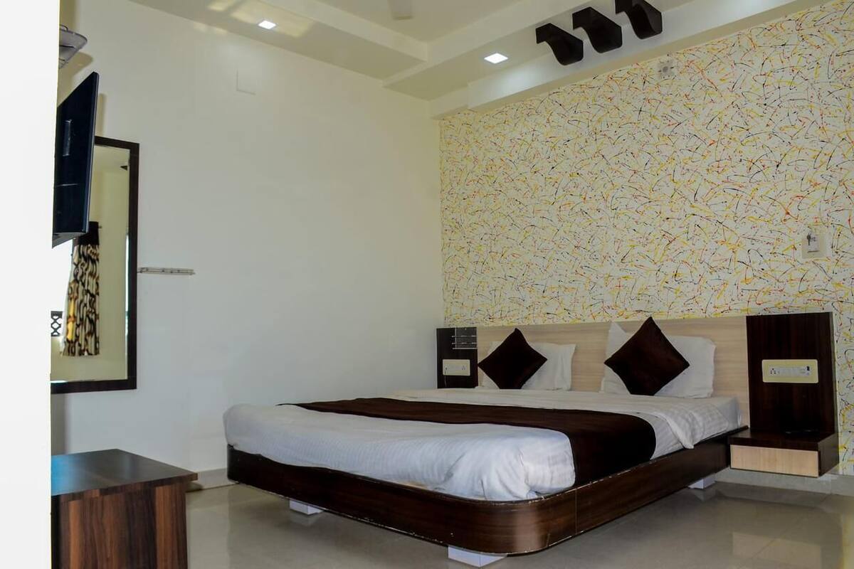A modern hotel room with a large bed, a flat-screen TV, a wooden nightstand, and a wall with a colorful abstract pattern.