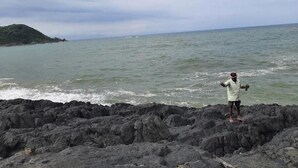 Une personne se tient sur un terrain rocheux, près d’une étendue d’eau, avec une masse terrestre visible au loin.
